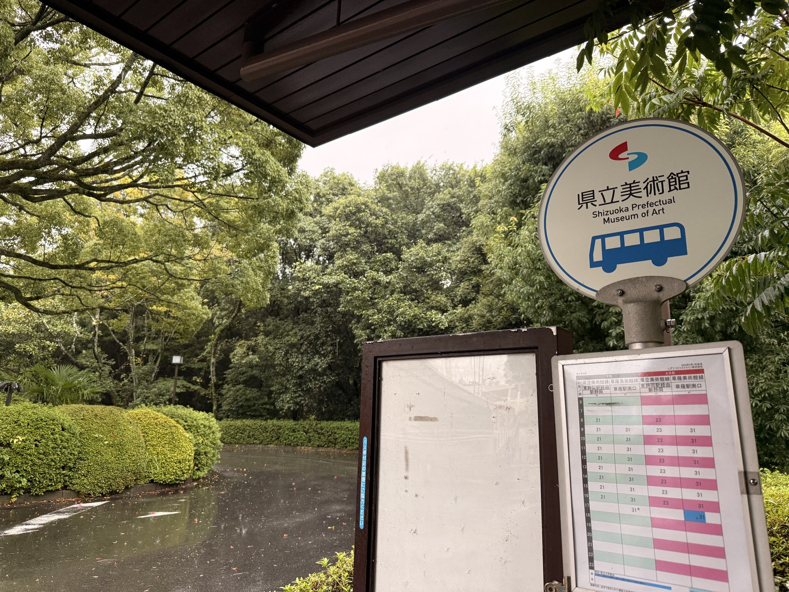 雨に濡れた木立の中に立つ「県立美術館」のバス停