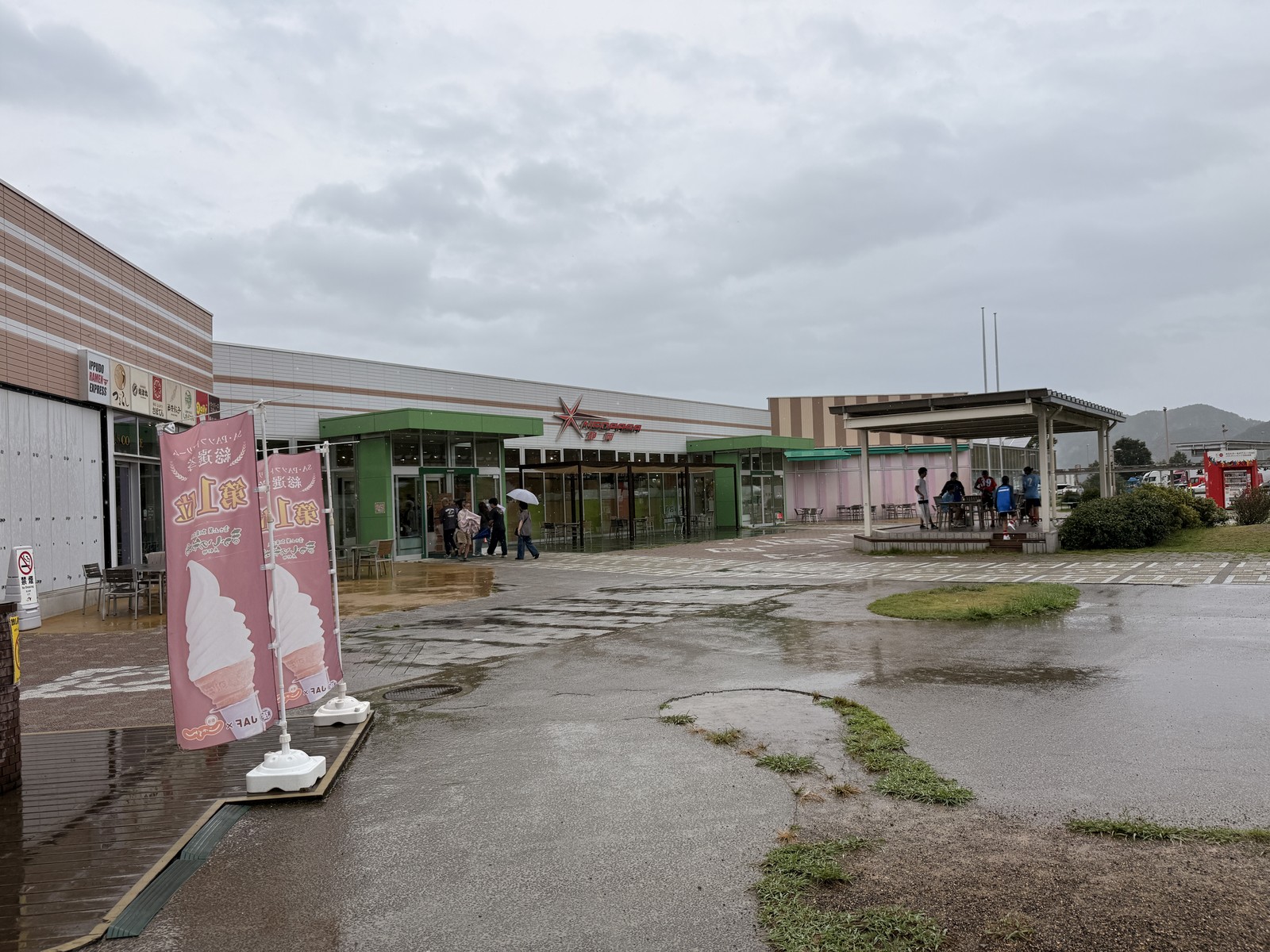 雨に濡れたNEOPASA静岡の駐車場、水たまりの向こうにソフトクリームののぼりが立っている